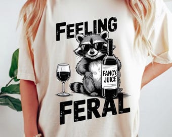 Op de afbeelding: Crèmekleurig T-shirt met een afbeelding van een wasbeer met zonnebril, die een wijnfles met het opschrift "Fancy Juice" en een wijnglas vasthoudt. De woorden "Feeling Feral" staan boven en onder de wasbeer.