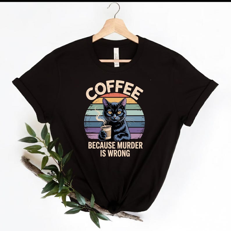 May include: Black t-shirt featuring a graphic of a black cat holding a coffee cup, with the text "COFFEE BECAUSE MURDER IS WRONG" in a circular design. The design includes a colorful sunset background.