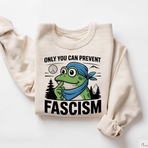 Puede incluir: Sudadera de color crema con una rana de dibujos animados que lleva un pañuelo azul y un dedo en los labios. El texto "ONLY YOU CAN PREVENT FASCISM" está impreso en negro debajo de la ilustración de la rana. La sudadera tiene mangas largas.