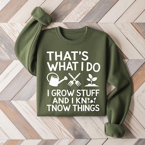 May include: An olive green sweatshirt with the text "THAT'S WHAT I DO I GROW STUFF AND I KNOW THINGS" in white. The design includes a watering can, crossed garden forks, and a sprouting plant. The sweatshirt has a crew neck and long sleeves.