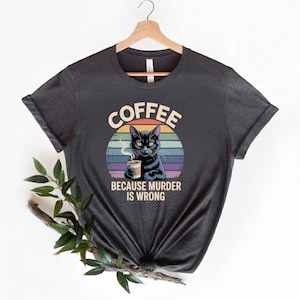 May include: Dark gray t-shirt featuring a black cat holding a coffee cup, set against a colorful sunset design. The text "COFFEE BECAUSE MURDER IS WRONG" is printed above and below the cat illustration.
