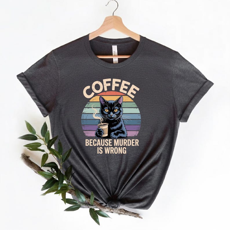 May include: Dark gray t-shirt featuring a black cat holding a coffee cup, set against a colorful sunset design. The text "COFFEE BECAUSE MURDER IS WRONG" is printed above and below the cat illustration.