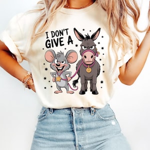 May include: Cream-colored t-shirt featuring a cartoon mouse and donkey with the text "I DON'T GIVE A". The mouse wears a star-shaped badge, and the donkey has a pink lead and collar. The design is surrounded by small black stars.
