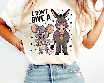 May include: Cream-colored t-shirt featuring a cartoon mouse and donkey with the text "I DON'T GIVE A". The mouse wears a star-shaped badge, and the donkey has a pink lead and collar. The design is surrounded by small black stars.