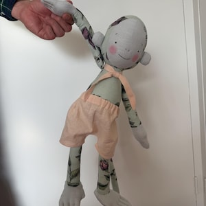 Può includere: Un giocattolo di scimmia di peluche fatto a mano con un corpo in tessuto a stampa floreale, pantaloncini color pesca e un viso grigio con guance rosa. La scimmia ha una coda lunga e sta tenendo fuori il braccio.