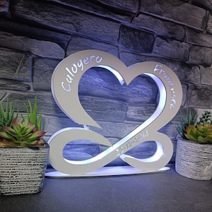 Può includere: Decorazione bianca a forma di cuore illuminata con i nomi "Calogero" e "Francesca" incisi su entrambi i lati. È visibile anche la data "23-12-2018". Il design incorpora un simbolo dell'infinito.