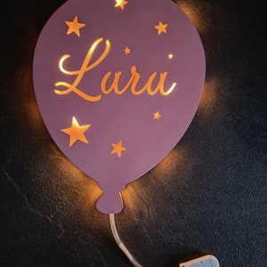 Puede incluir: Una luz nocturna en forma de globo de color morado con el nombre "Lara" recortado e iluminado con luces LED blancas cálidas. El globo está decorado con pequeñas estrellas doradas.