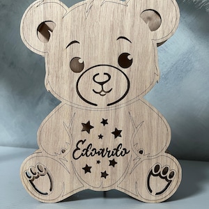 Può includere: Sagoma di legno di un orso con un viso sorridente. L'orso è seduto con le zampe distese. L'orso ha un disegno a stella sul petto con il nome "Edoardo" scritto al suo interno.