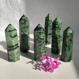 May include: Seven green, black, and pink zoisite crystal points arranged in a row on a white surface.  Pink flowers are in the foreground.
