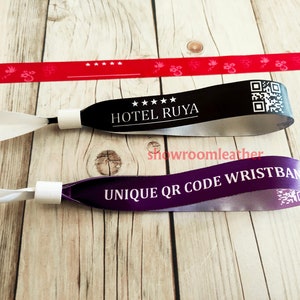 Op de afbeelding: Twee polsbandjes met unieke QR-codes. Het zwarte polsbandje heeft de tekst "HOTEL RUYA" met vijf sterren. Het paarse polsbandje heeft de tekst "UNIQUE QR CODE WRISTBAND".