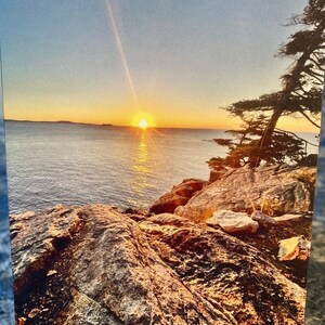 May include: A scenic sunset over a body of water with rocky cliffs in the foreground. The sun is setting behind the horizon, casting a golden glow over the water and rocks.