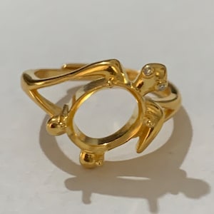 May include: A gold-colored ring in the shape of a sea turtle. The ring features a circular setting for a gemstone and small, sparkling accents. The turtle design includes the head, flippers, and shell, all crafted in a shiny gold metal.