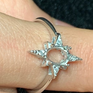 May include: A silver ring with a starburst design and a large oval setting for a gemstone. The ring is set with small clear stones around the starburst and the oval setting.