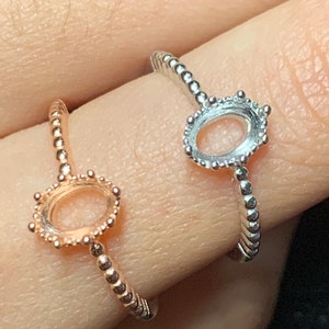 May include: Two oval-shaped ring settings, one in rose gold and the other in silver, are displayed. The bands feature a beaded design, and the settings are designed to hold gemstones. The rings are shown on a finger.