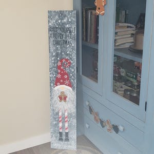 May include: A wooden sign featuring a painted gnome. The gnome is wearing a red hat with white snowflakes and a red and white striped scarf. The sign reads "Have yourself a merry little Christmas".
