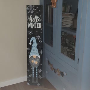 Könnte beinhalten: Ein Holzschild mit einem Zwerg und dem Text "hello winter" darauf. Das Schild ist mit Schneeflocken verziert und in Schwarz, Weiß und Blau bemalt.