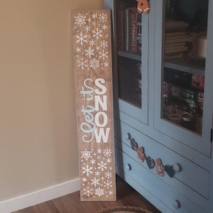Op de afbeelding: Een houten bord met de woorden "Let it Snow" in witte en lichtblauwe letters. Het bord is versierd met witte sneeuwvlokken en heeft een rustieke, verweerde uitstraling. Het bord leunt tegen een muur naast een blauwe kast.
