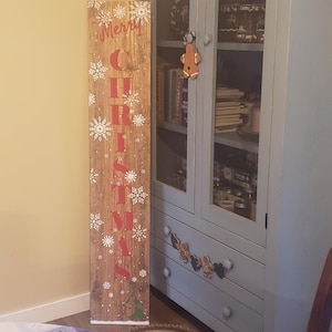 May include: A tall, rectangular wooden porch sign with a faux wood grain finish. The sign features the word "CHRISTMAS" in large, vertical red capital letters, accented by white snowflake illustrations and the word "Merry" at the top.