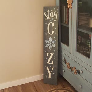 May include: A grey wooden sign with white lettering that reads "Stay Cozy" with a snowflake design. The sign is leaning against a light blue cabinet.