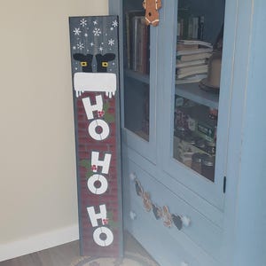 Könnte beinhalten: Ein langes, rechteckiges Weihnachtsschild mit Santa Claus-Motiv. Oben sind die Stiefel des Weihnachtsmanns und Schneeflocken zu sehen. Auf dem Schild steht "HO HO HO" in weißen Buchstaben auf rotem Ziegelgrund. Im Hintergrund ein blauer Schrank.
