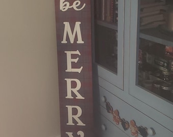 Puede incluir: Un letrero de madera con las palabras "be merry" pintadas en blanco sobre un fondo de cuadros rojos y negros.