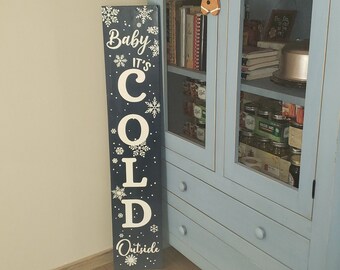 Può includere: Un cartello di legno blu con lettere bianche che recita "Baby It's Cold Outside". Il cartello è decorato con fiocchi di neve bianchi.