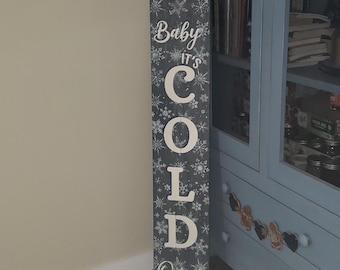 Puede incluir: Un letrero de madera rectangular y alto con un fondo gris oscuro y diseños de copos de nieve blancos. El letrero dice "Baby It's Cold Outside" en letras blancas, perfecto para la decoración de invierno. El letrero es probablemente para uso en interiores o exteriores.