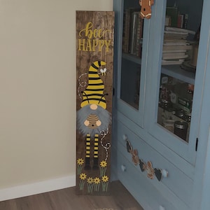 Op de afbeelding: Een houten bord met een bij-thema dwerg en de woorden "be happy" erop geschilderd. De dwerg draagt een zwart-gele gestreepte hoed en heeft een bij op zijn schouder.