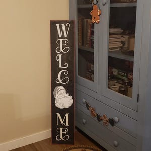 Könnte beinhalten: Ein dunkelbraunes Holz-Willkommensschild mit weißer Schrift und einer Illustration des Weihnachtsmanns. Das Schild ist vertikal und steht neben einem blauen Schrank mit Glastüren. Der Schrank ist mit verschiedenen Gegenständen gefüllt.