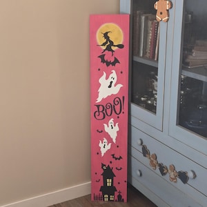 May include: A tall, rectangular pink wooden sign featuring a painted Halloween scene. The design includes a yellow full moon, a black silhouette of a witch on a broomstick, white ghosts, black bats, the word BOO in black, and a black haunted house.