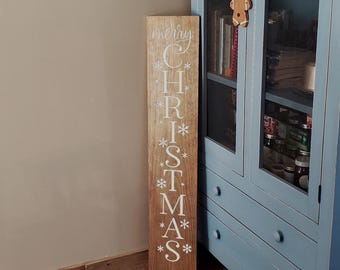 Puede incluir: Un letrero de madera alto y rectangular con un acabado natural en color marrón claro. La palabra "CHRISTMAS" está impresa verticalmente en letras mayúsculas blancas, con la palabra "merry" en cursiva blanca arriba y copos de nieve blancos.