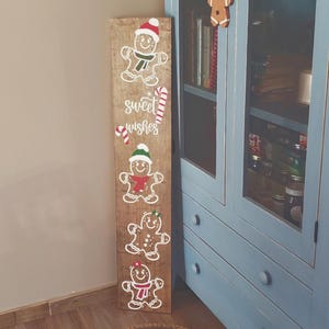 May include: A tall, rectangular wooden sign with a distressed finish featuring four white gingerbread man illustrations, each wearing a different coloured scarf and hat, with the text "sweet wishes" and two candy cane graphics in the centre.