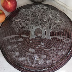 May include: A round, clear glass plate with an etched landscape design featuring trees, flowers, and a small bridge. The plate has a dark brown woven border. Two red apples are visible in the upper left corner.