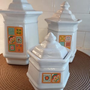 May include: Three white ceramic canisters with lids, each featuring a colorful square design with floral and geometric patterns. The canisters are arranged on a brown woven placemat, showcasing a vintage kitchenware set.