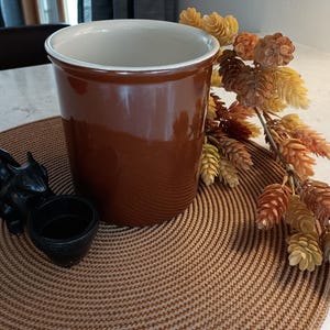 May include: A brown ceramic utensil holder with a white interior rim sits on a brown woven placemat. A black decorative elephant-shaped candle holder and a branch of faux fall foliage complete the arrangement.