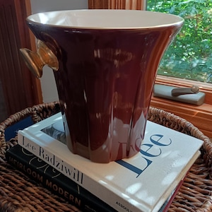 May include: A deep brown, fluted ceramic ice bucket with a smooth, glossy finish and a cream-colored interior. The vessel features two gold-toned, knob-shaped side handles and sits atop a stack of two hardcover books, Lee Radziwill and Heirloom Modern.