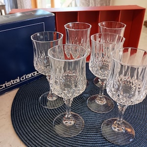 May include: Six clear crystal wine glasses with a diamond pattern, arranged on a blue placemat. A blue box with the text "cristal d'arques" is visible in the background, along with a red box.
