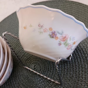 May include: White porcelain serving bowl with a scalloped blue rim and floral pattern. The bowl is supported by a twisted metal stand. A stack of matching plates is visible to the left. The bowl is approximately 6 inches wide.