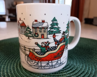 May include: White ceramic mug with a winter scene illustration. The design features a red sleigh with two people, houses, and evergreen trees. The mug has a curved handle and is set on a green textured surface.