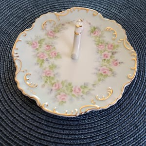May include: A two-tiered serving plate with a light blue rim and a white center. The plate is decorated with pink roses and green leaves. The rim is trimmed with gold. A white handle with gold accents connects the two tiers.