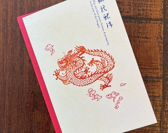 May include: A red and orange dragon illustration on a white card with a red border. The card has the text "May you have peace, fortune, & prosperity in the year of the Dragon."