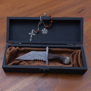 Puede incluir: Una caja de madera negra con un forro de terciopelo marrón. La caja contiene un cuchillo plateado con mango negro y la palabra "THAMSPHERE" inscrita en la hoja. También se puede ver una cruz plateada y un pentagrama en la caja.