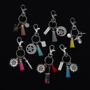 Könnte beinhalten: Satz von 8 silbernen Schlüsselanhängern mit verschiedenen Anhängern, darunter ein Pentagramm, ein Messer, ein Schädel und ein Quaste. Die Anhänger sind an silbernen Schlüsselringen mit silbernen Clips befestigt.