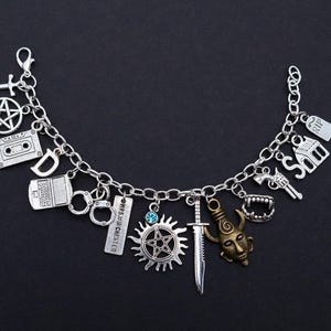 May include: Silver charm bracelet with a variety of pendants, such as a pentagram, cassette tape, handcuffs, and a sword. Other charms include a house, a gun, and a devil's head. The bracelet is displayed against a black backdrop.