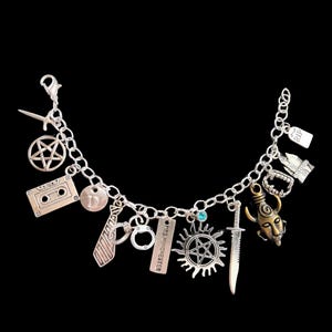 Puede incluir: Pulsera de abalorios plateada con varios dijes, incluyendo un pentagrama, una cinta de cassette, una corbata, esposas y una espada. Otros dijes incluyen un diseño de sol, una cara y una lápida.