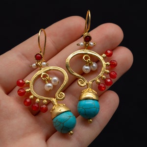 Op de afbeelding: Goudkleurige oorbellen met turquoise, rode en witte accenten. De oorbellen hebben een druppelvorm met een turkooizen kraal aan de onderkant, rode kralen en witte parels. De oorbellen worden vastgehouden door een gouden haak.