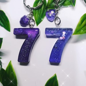 May include: Purple glitter resin keychain featuring the numbers 77. The numbers are attached to a silver chain with a small purple heart charm.