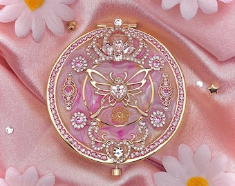 Könnte beinhalten: Ein goldfarbener Kompaktspiegel mit Schmetterlingsdesign, besetzt mit rosa Strasssteinen und klaren Kristallen. Der Spiegel hat Kronen- und Herzverzierungen, vor einem rosa-weißen Marmorhintergrund. Rosa Gänseblümchen umgeben den Kompaktspiegel.