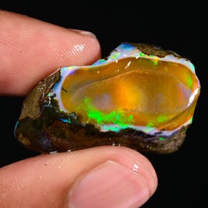 May include: A rough opal gemstone with a vibrant display of colors. The opal features a golden-yellow interior with flashes of green, blue, and orange. The stone is held between fingers, showcasing its natural, raw form. The opal's surface is rough and textured.
