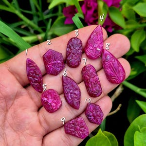 May include: Ten individual deep pink, translucent gemstones, each carved with a floral pattern and labelled with a white number from one to ten. The stones vary in shape, including teardrop, oval, and geometric forms, displaying a polished finish.
